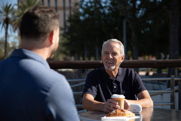 Two men having an outdoor breakfast, one of them wearing Explore Li+ has placed charging case on the table