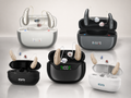 Five pairs of behind-the-ear hearing aids with their open charging cases arranged on a light gray reflective surface. The hearing aids come in beige, white, and dark gray/black colorways, with digital battery levels visible on the cases.